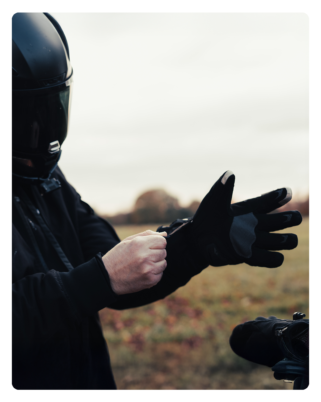 A rider pulling on a heated glove in an open field, the other waiting on the bike.
